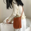 A close-up of an elegant pleated faux leather crossbody bag in a warm caramel brown color resting on a white surface.