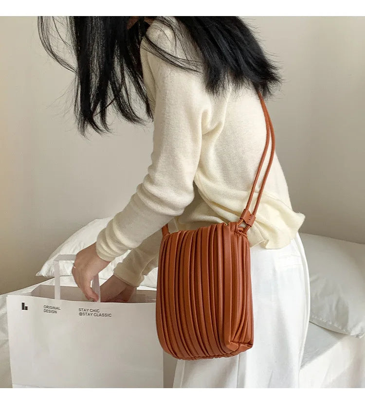 A close-up of an elegant pleated faux leather crossbody bag in a warm caramel brown color resting on a white surface.