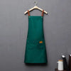 A dark green waterproof kitchen apron hanging on a wooden hanger against a gray wall.
