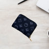 A dark navy zippered card holder wallet with a repeating geometric pattern rests on a light wooden surface next to a pen and 