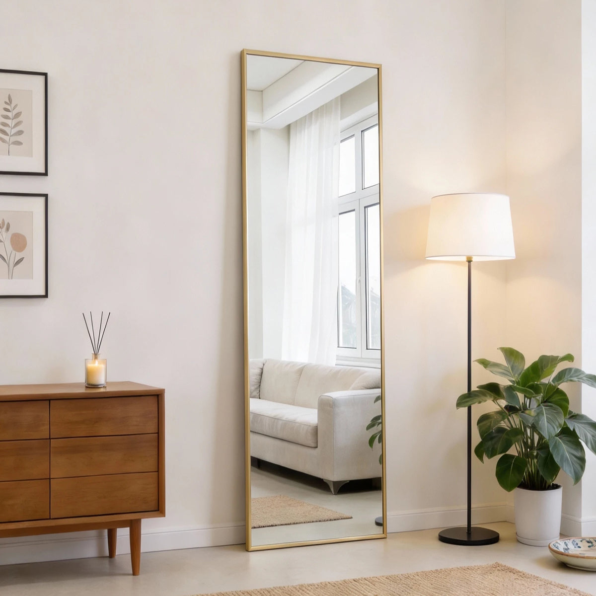 A full length rectangular mirror with a gold frame leaning against a white wall in a bright room.