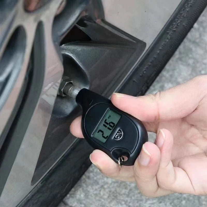 A hand holds a black digital tire pressure gauge with an LCD display near a car wheel.