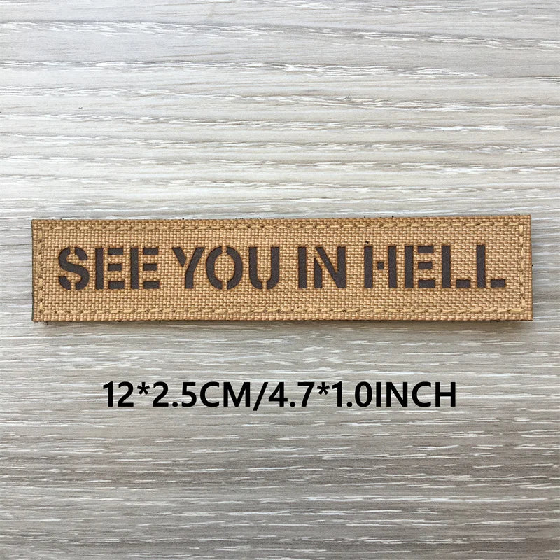 A rectangular tan woven patch with black text that reads SEE YOU IN HELL on a light wooden surface.