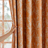 An orange curtain panel with a white plant pattern is being drawn open by a hand near a window.
