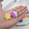 A hand holds a purple and white squishy cat paw fidget toy and a yellow squishy cat paw fidget toy.