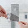 A hand places a self-adhesive fiberglass screen repair patch onto a mesh surface.