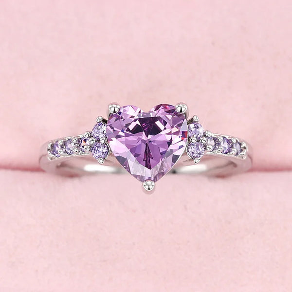 A dainty heart ring featuring a large purple gemstone surrounded by smaller cubic zirconia stones.