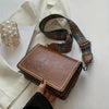 A brown leather mini cross-body bag with an adjustable patterned strap is shown next to a cream-colored shirt.