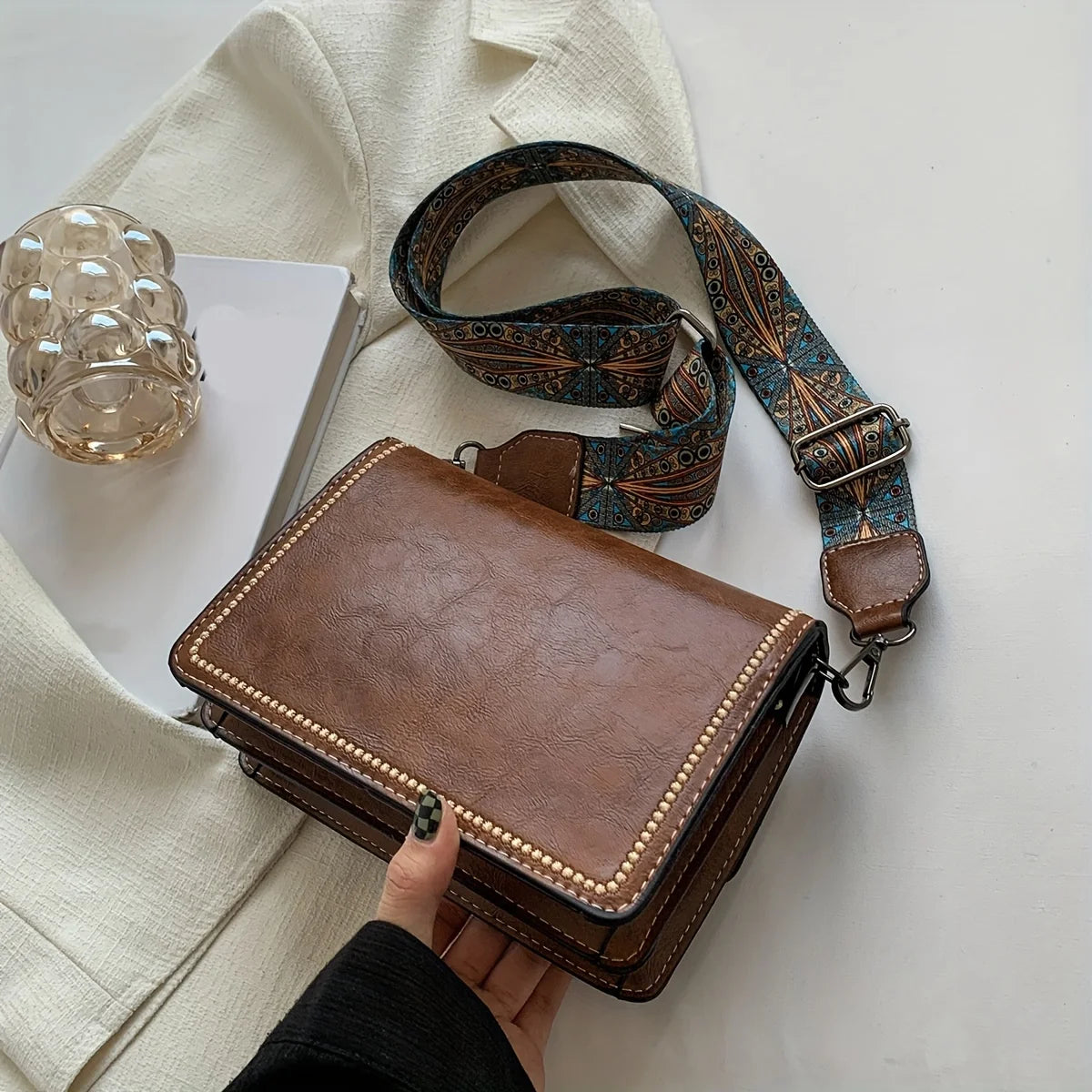 A brown leather mini cross-body bag with an adjustable patterned strap is shown next to a cream-colored shirt.