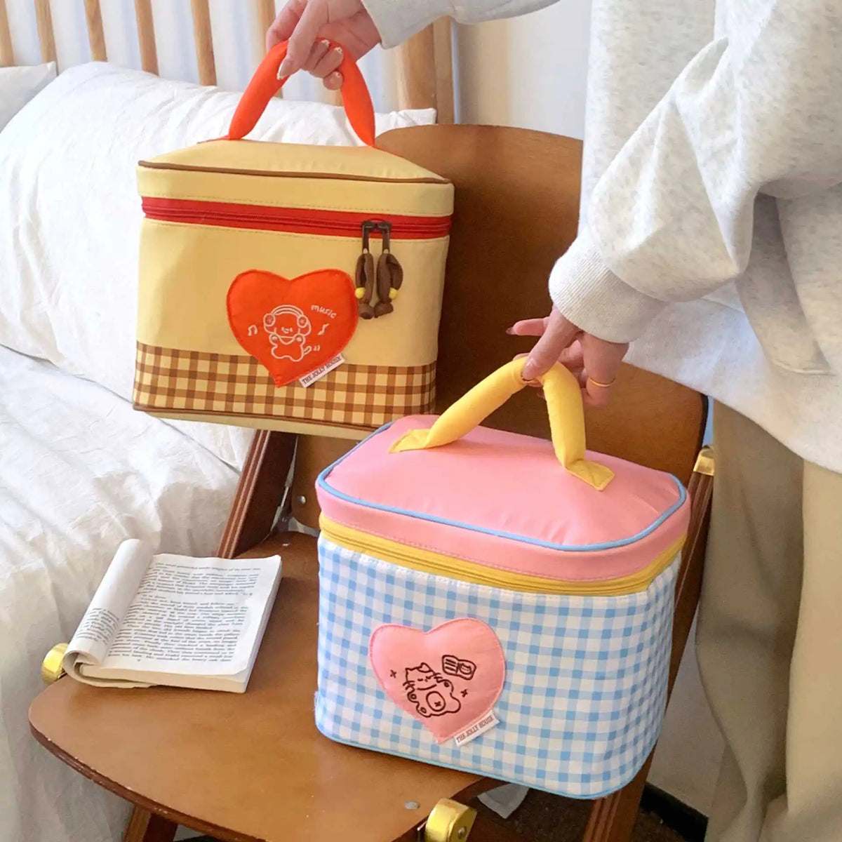 A hand is holding a yellow insulated lunch bag with a cartoon design, next to a pink one.