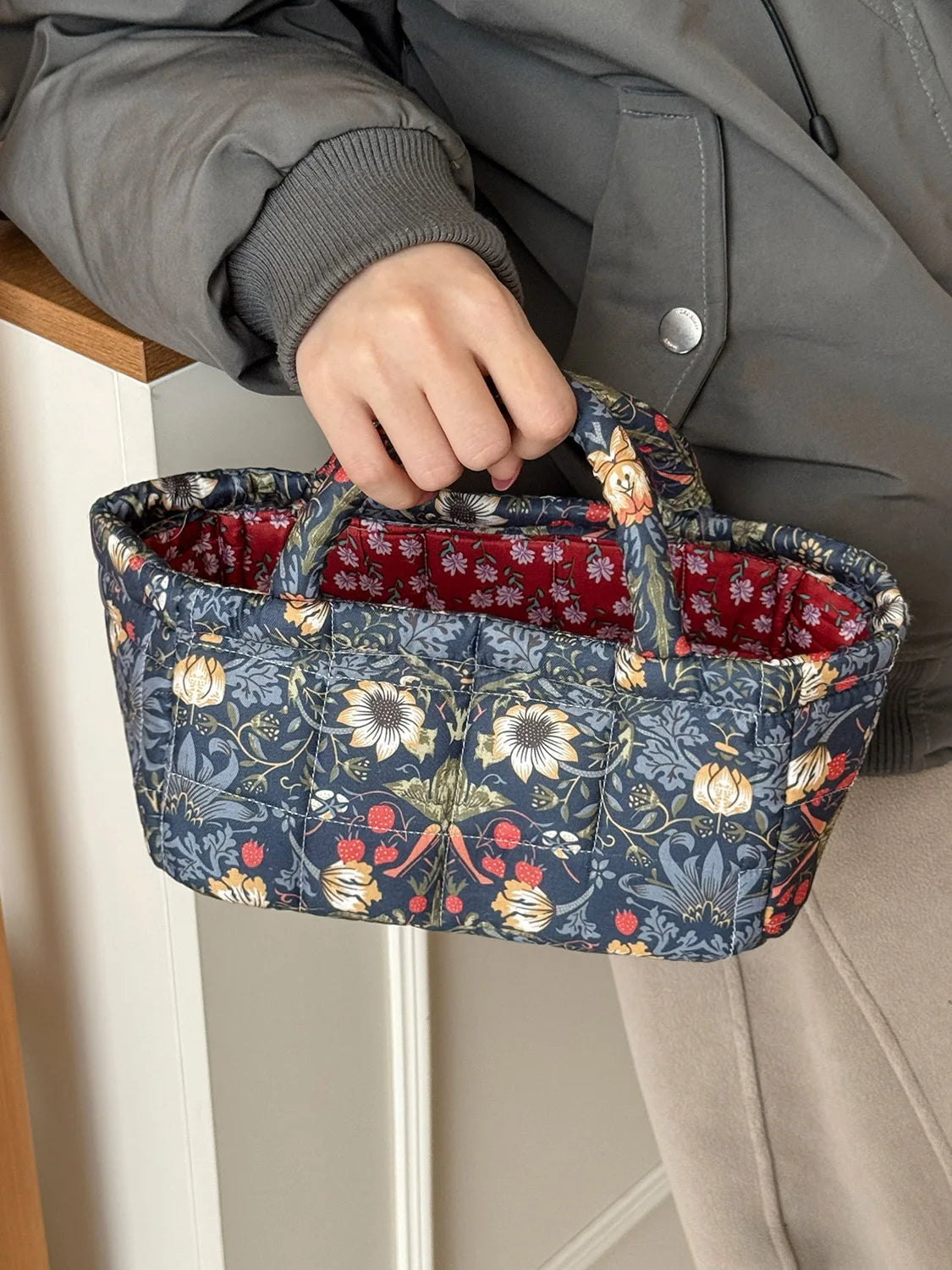 A hand holds a quilted fabric storage basket with a blue floral pattern, featuring a red lining, against a light background.