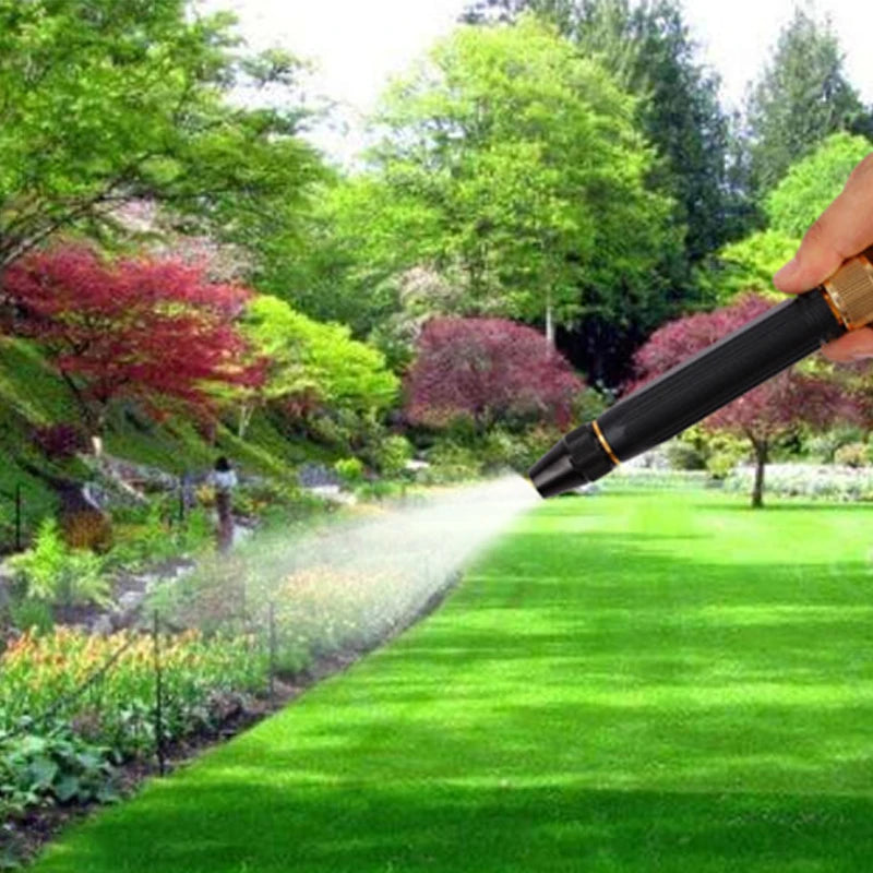 A hand is holding an adjustable high-pressure water gun spraying a fine mist onto a vibrant green lawn surrounded by lush tre