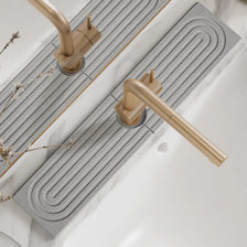 A brass faucet mat with a wavy design is installed on a white countertop next to a kitchen sink.