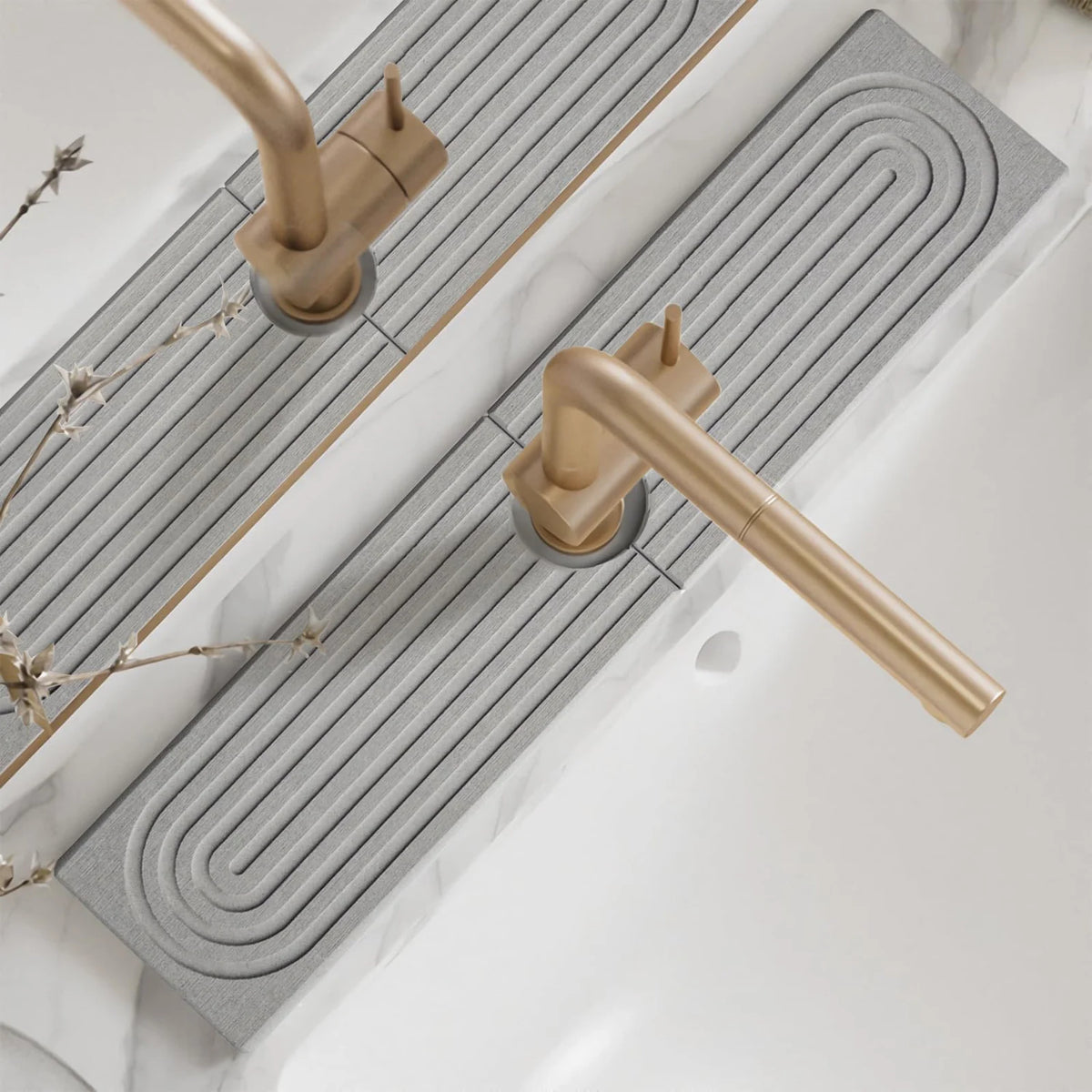 A brass faucet mat with a wavy design is installed on a white countertop next to a kitchen sink.