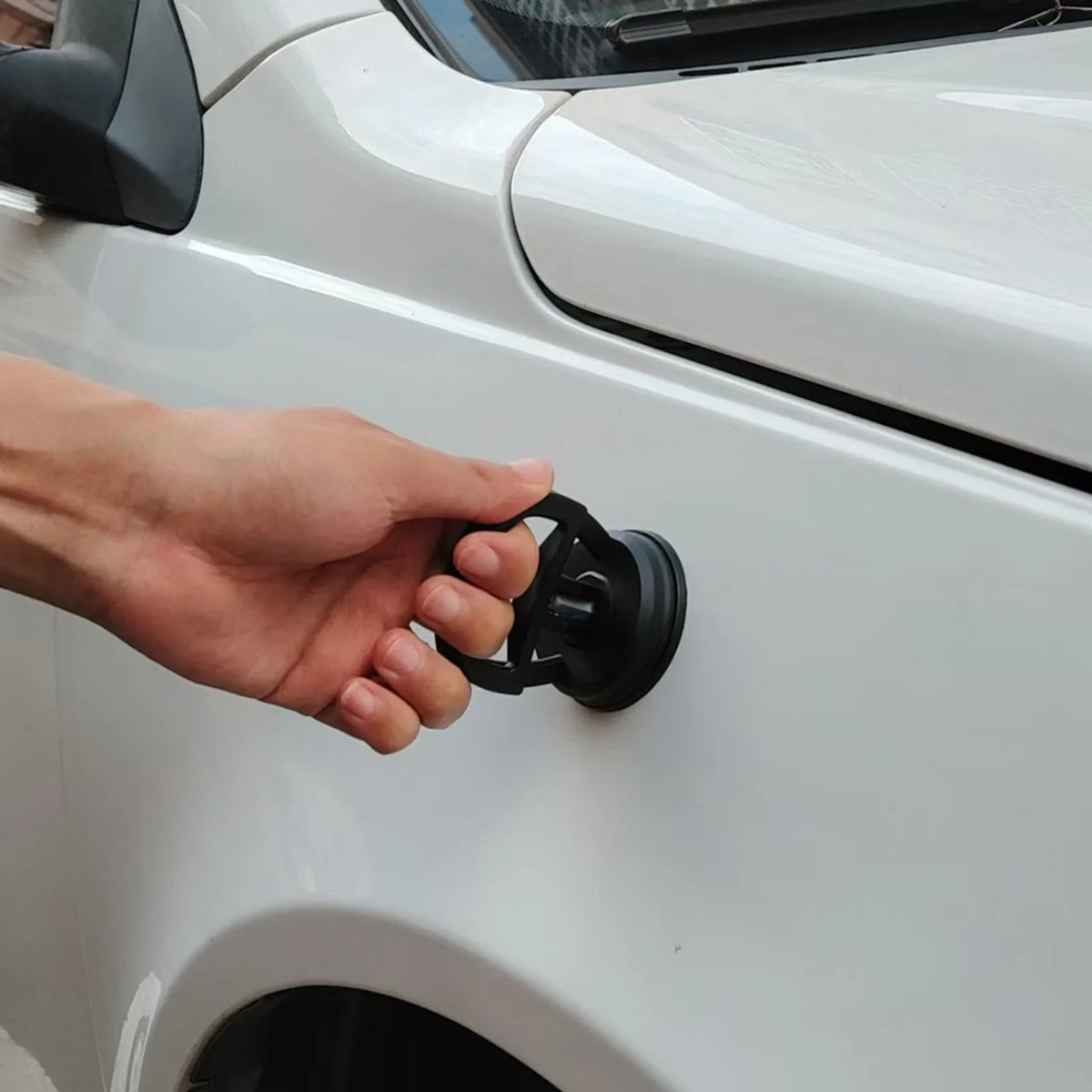 A hand is operating the handle of a car dent puller kit, which is attached to the side of a white car.