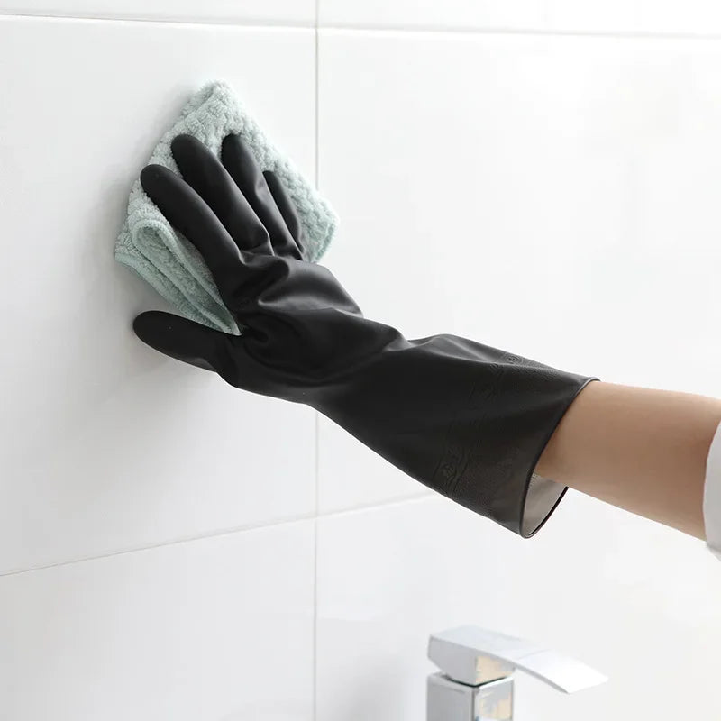 A hand wearing a black rubber cleaning glove is wiping a white tile with a green textured cleaning cloth.