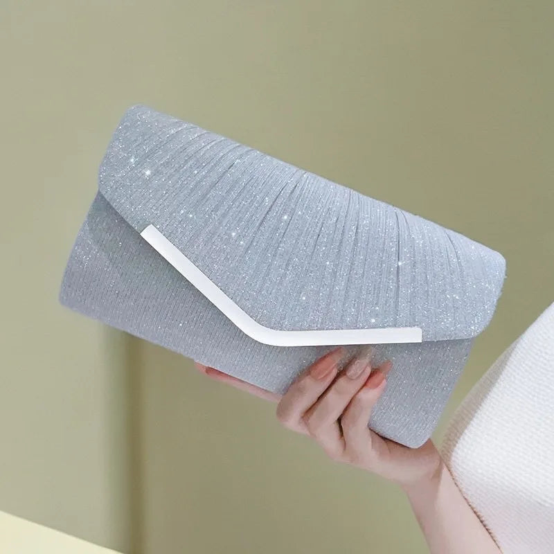 A light gray pleated glitter envelope clutch with a white strap is held by a hand.