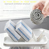 A hand holds a coiled gray cleaning sponge, next to two blue and white cleaning sponges resting in a stainless steel holder o