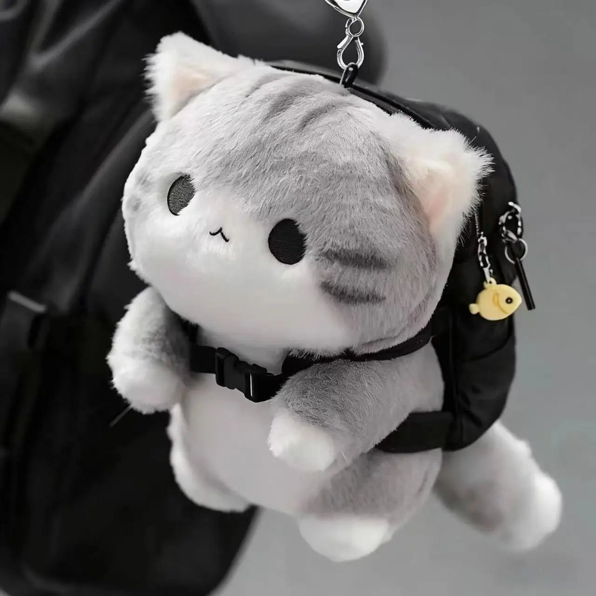 A soft grey cat plush toy with black backpack charm hanging from a black strap.