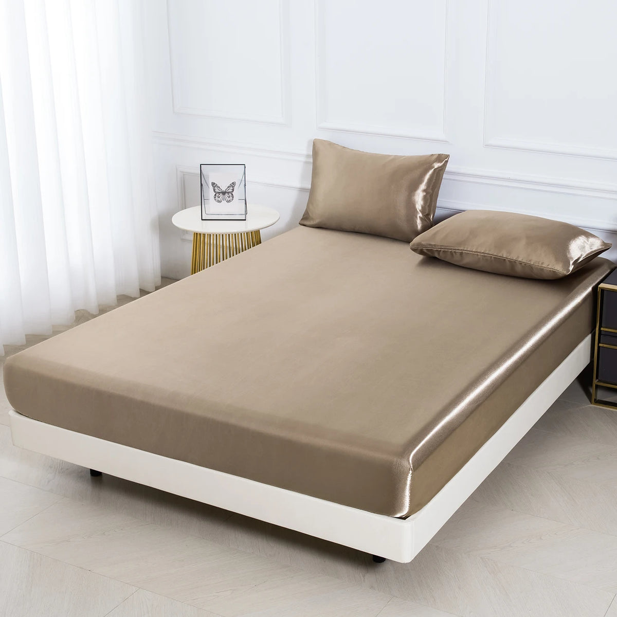 Beige Satin-Bettwäsche-Set mit zwei passenden Kissenbezügen auf einem weißen Bettrahmen in einem hellen Raum.