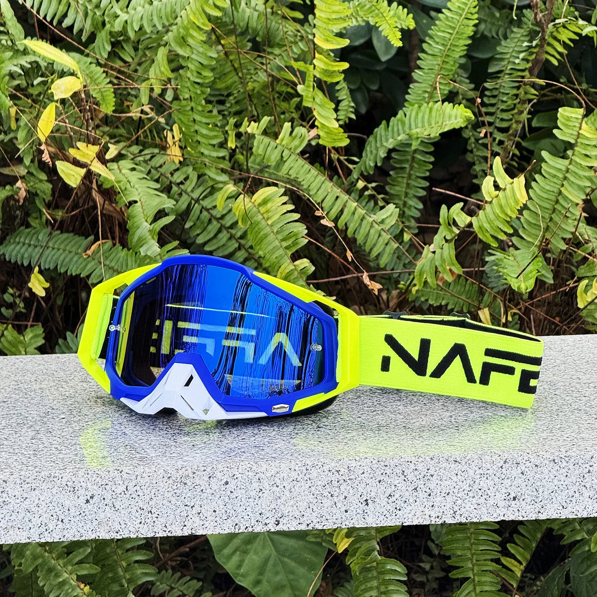 A pair of blue and yellow NAFE motocross goggles resting on a gray surface amidst green foliage.