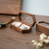 A wooden ring box with an open lid displaying several rings and a velvet pouch on a dark wood surface.