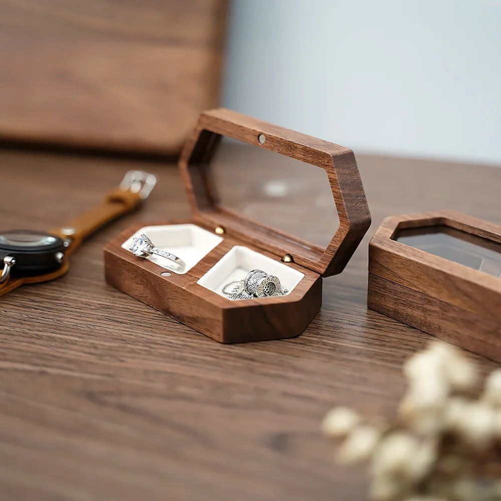 A wooden ring box with an open lid displaying several rings and a velvet pouch on a dark wood surface.