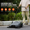 A person is pushing a black walk-behind push floor sweeper across an outdoor paved surface.