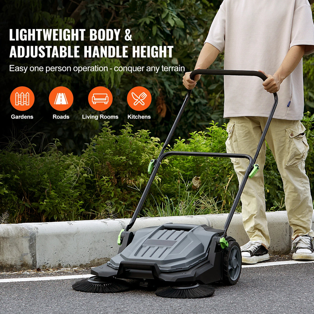 A person is pushing a black walk-behind push floor sweeper across an outdoor paved surface.