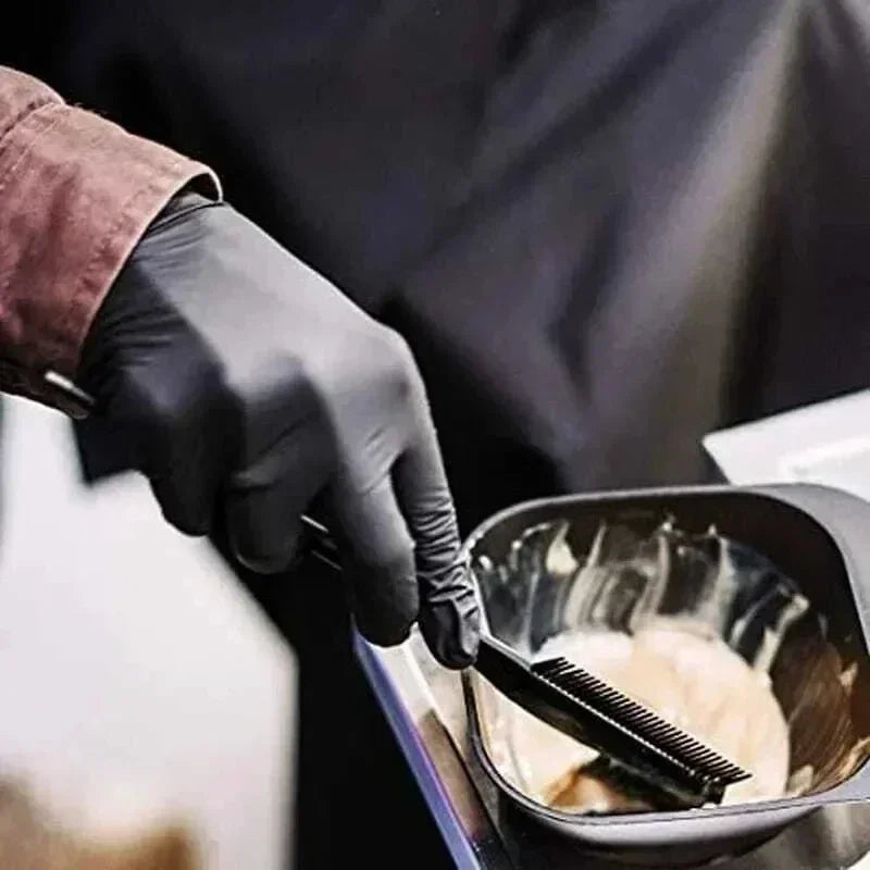 A hand wearing a black nitrile disposable glove is using a brush to apply a light-colored substance in a dark bowl.