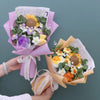 Sträußchen aus handgefertigtem Häkelblumen und verschiedenen Blumen in Gelb-, Violett- und Grüntönen, in rosa Papier eingewic