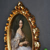 An ornate gold-framed portrait of a woman with dark hair and a white dress.