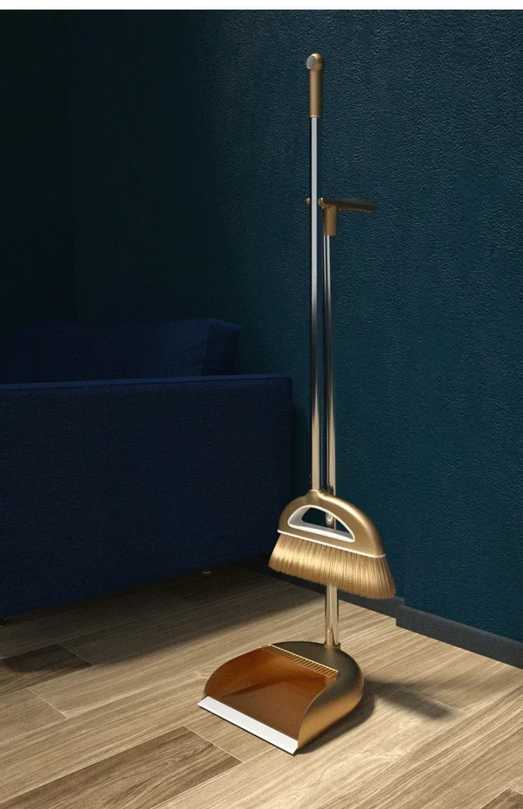 A modern brass broom and dustpan set with a floor wiper resting on a wooden floor.