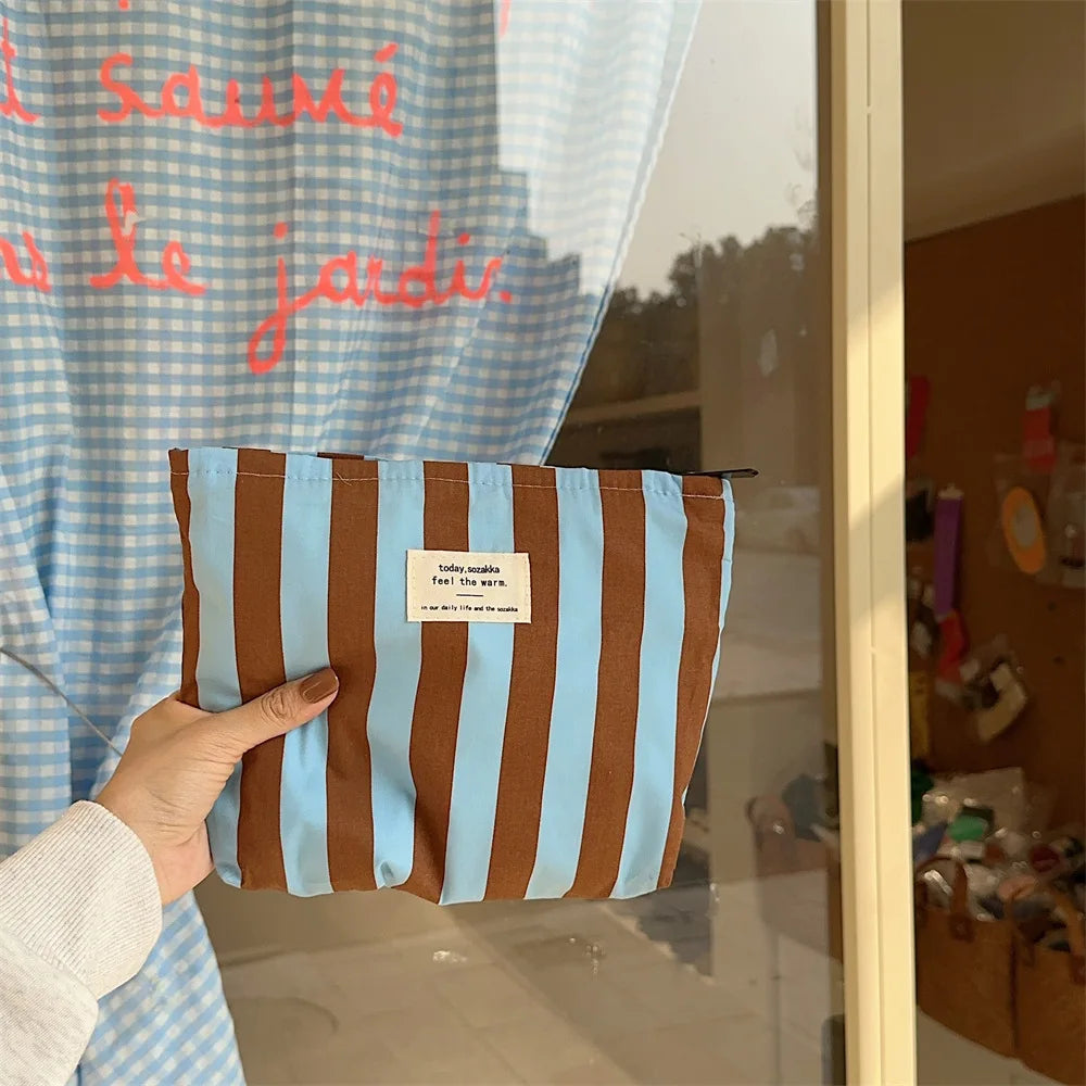A brown and light blue striped canvas cosmetic bag is held in a person's hand against a glass background.