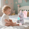 A baby is sitting on a bed with a gray elephant plush toy and a pink bow.