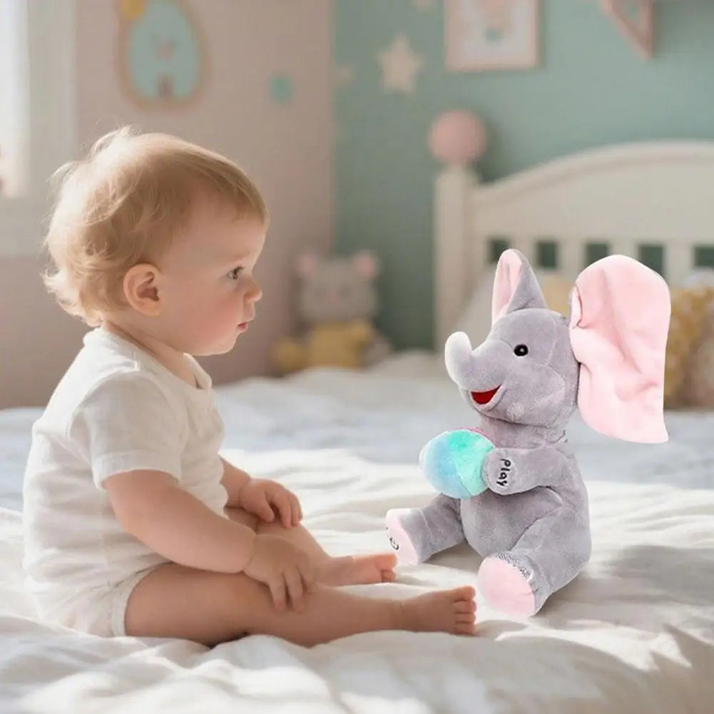 A baby is sitting on a bed with a gray elephant plush toy and a pink bow.
