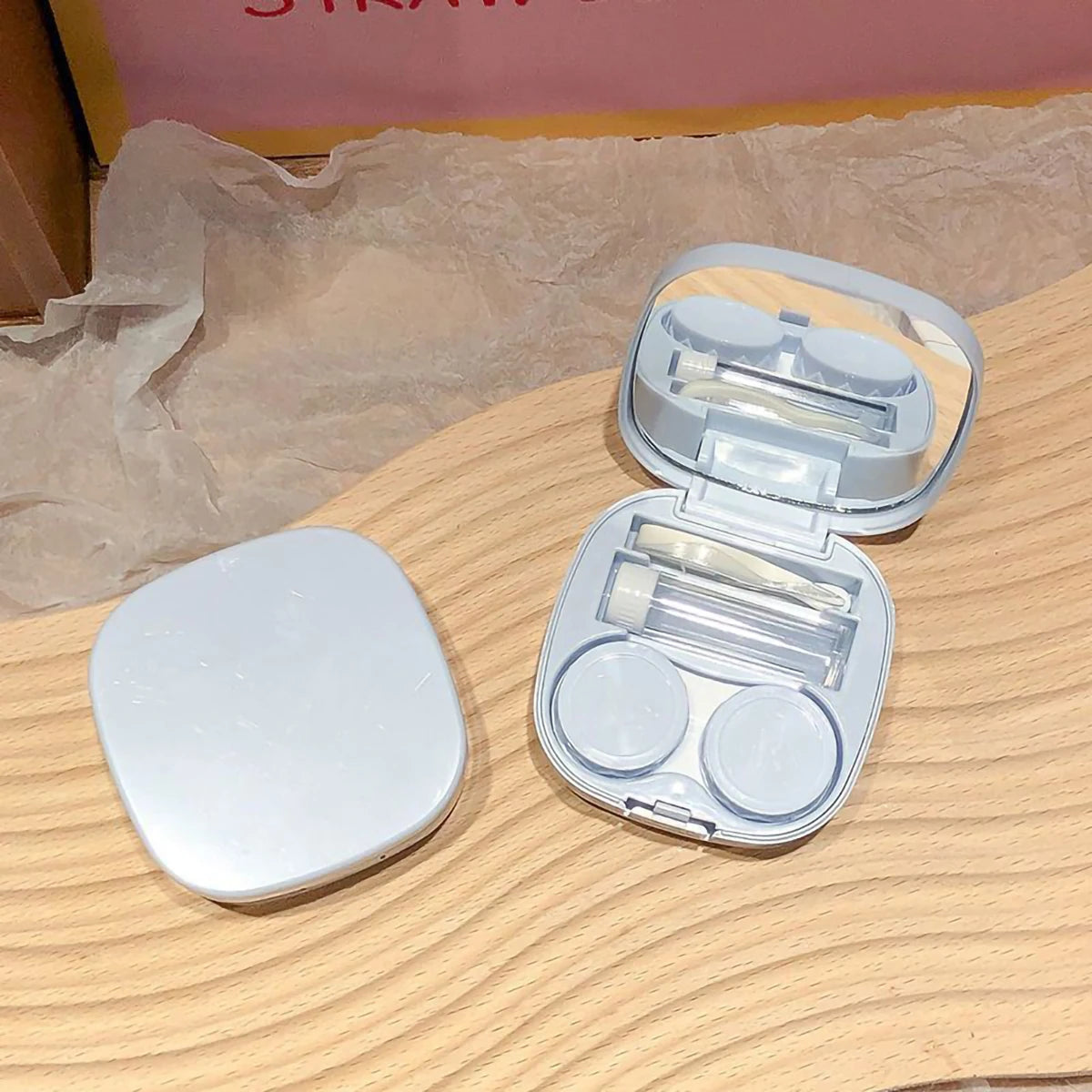 An open compact contact lens travel case showing clear lenses and storage compartments on a wooden surface.