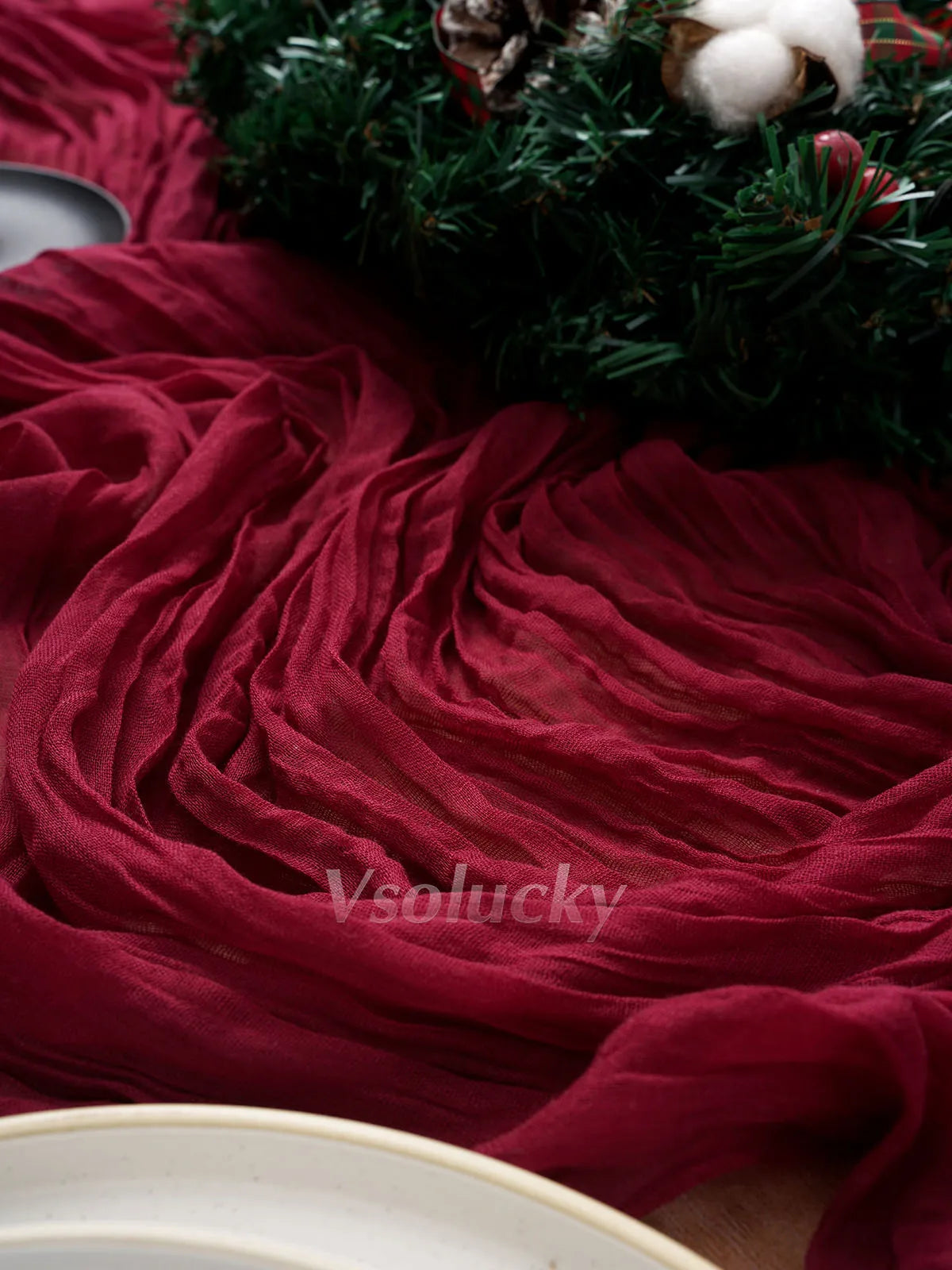 A deep red crinkle gauze cheesecloth table runner draped next to a green festive wreath.