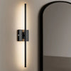 A black rechargeable LED wall sconce with a long vertical arm is mounted on a beige wall next to a large mirror.