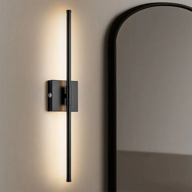 A black rechargeable LED wall sconce with a long vertical arm is mounted on a beige wall next to a large mirror.