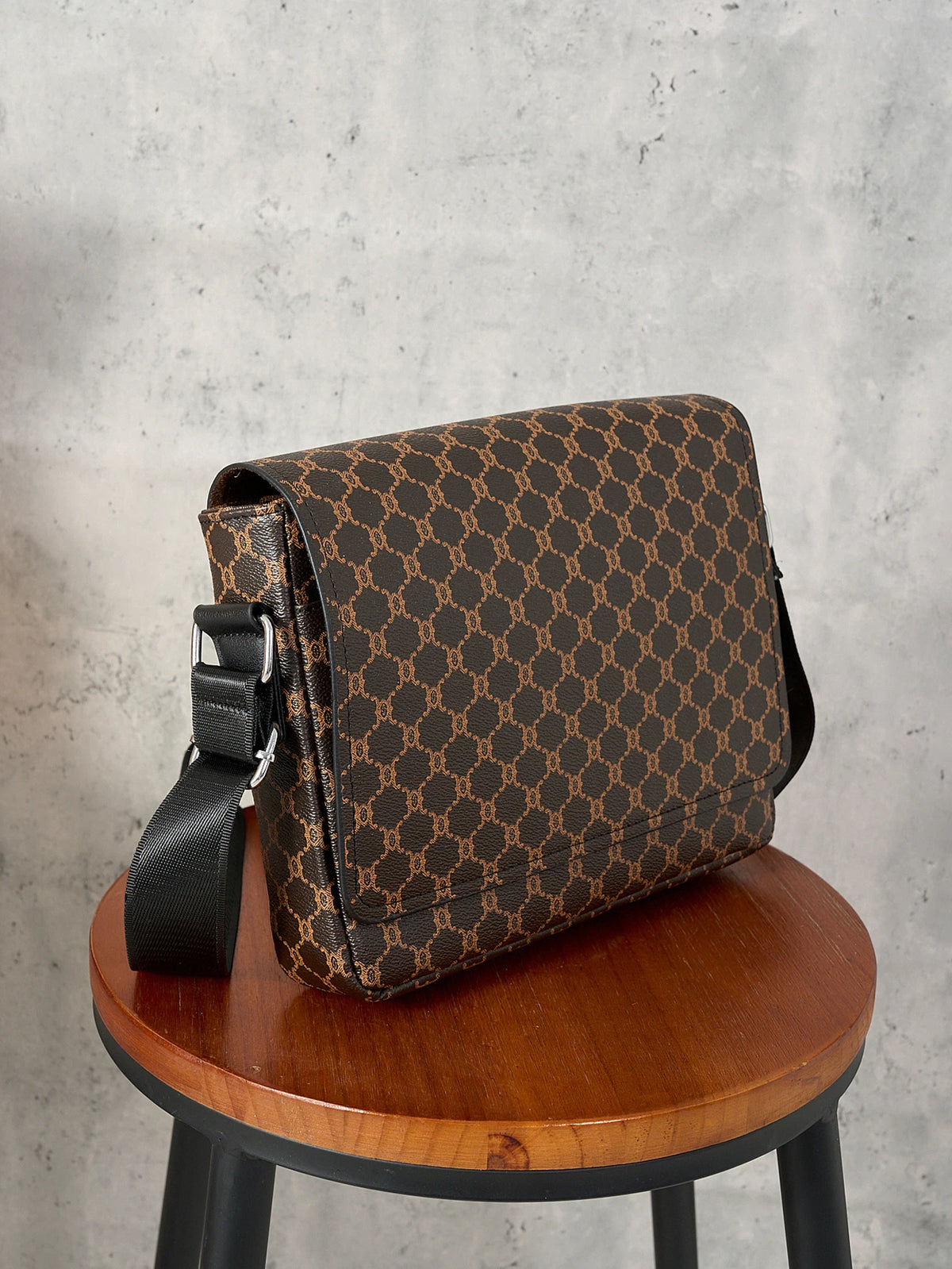 A brown patterned crossbody bag with a black strap resting on a wooden table against a gray background.