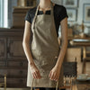 A person wearing a beige cotton apron over a black shirt stands in a rustic kitchen setting.