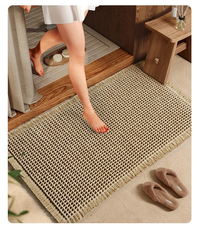 A close-up of a beige waffle weave bath mat with tassels placed on a wooden floor.
