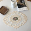 An elegant oval lace embroidered placemat with a floral design is displayed on a white table next to a ceramic container and 