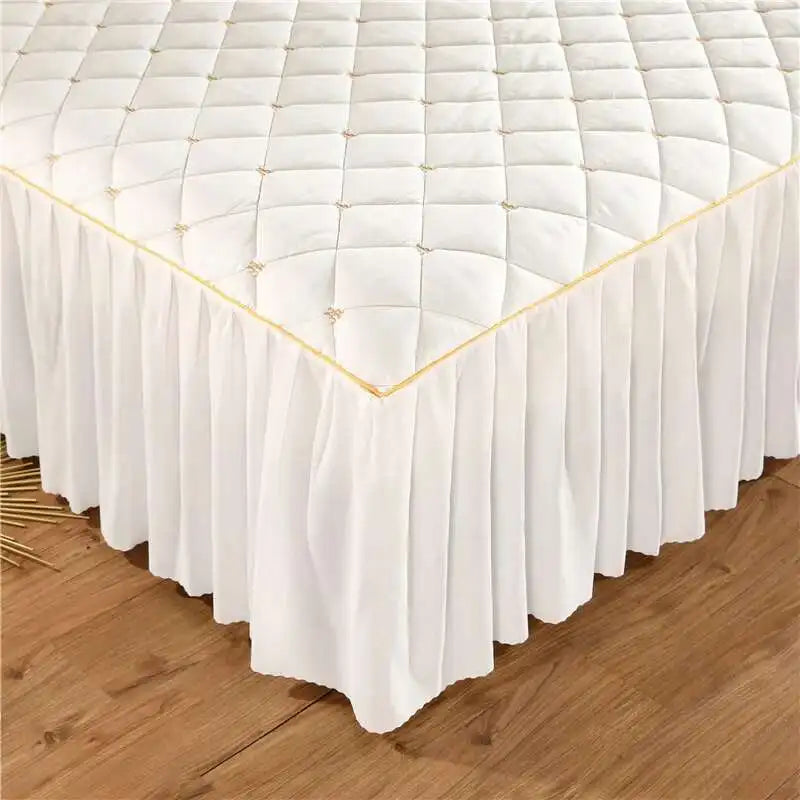 White quilted embroidered bed skirt with gold trim draped on a wooden floor.