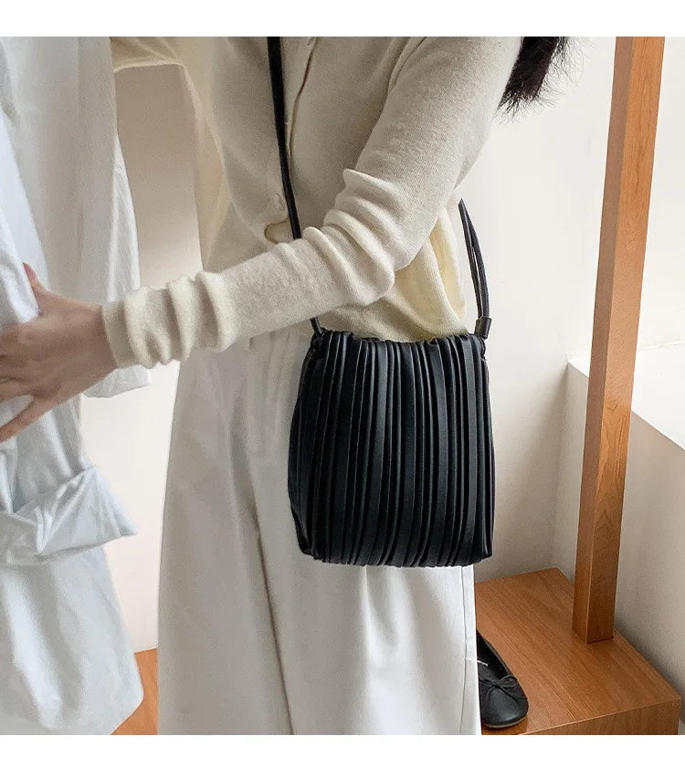 A woman wearing a white dress holds an elegant black pleated faux leather crossbody bag.