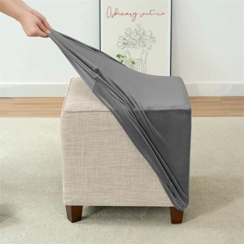 A gray stretch velvet ottoman slipcover is being placed over a light beige upholstered square pouf.