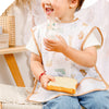 A smiling baby wearing a white feeding bib and blue jeans, holding a glass of water.
