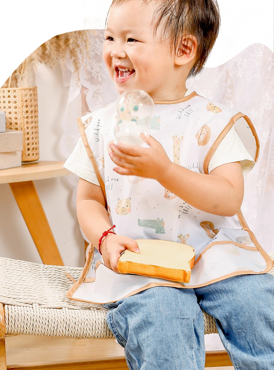 A smiling baby wearing a white feeding bib and blue jeans, holding a glass of water.