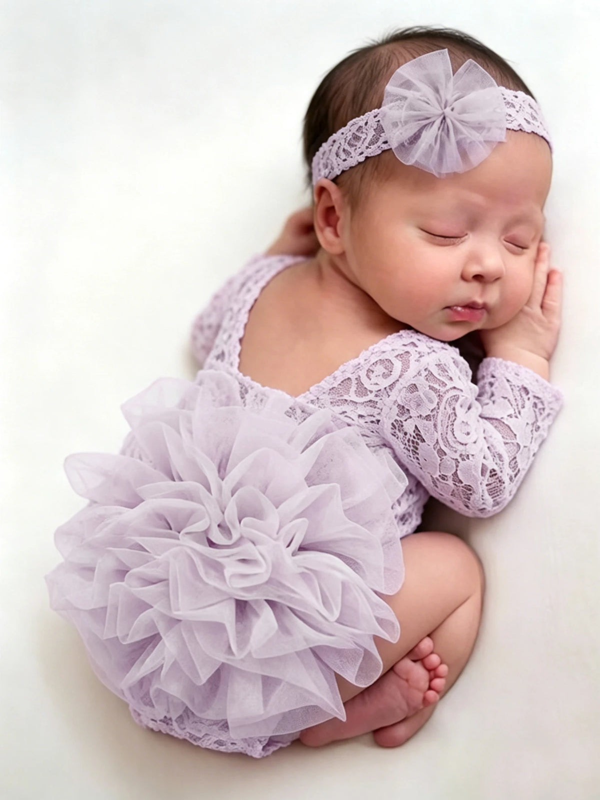 A newborn baby wearing a lavender lace romper with a ruffled tutu and a matching headband, lying down.
